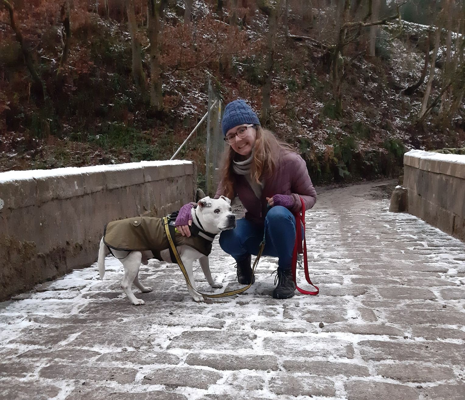 Martha and her white dog look at the camera on a snowy cobbled bridge in a wood